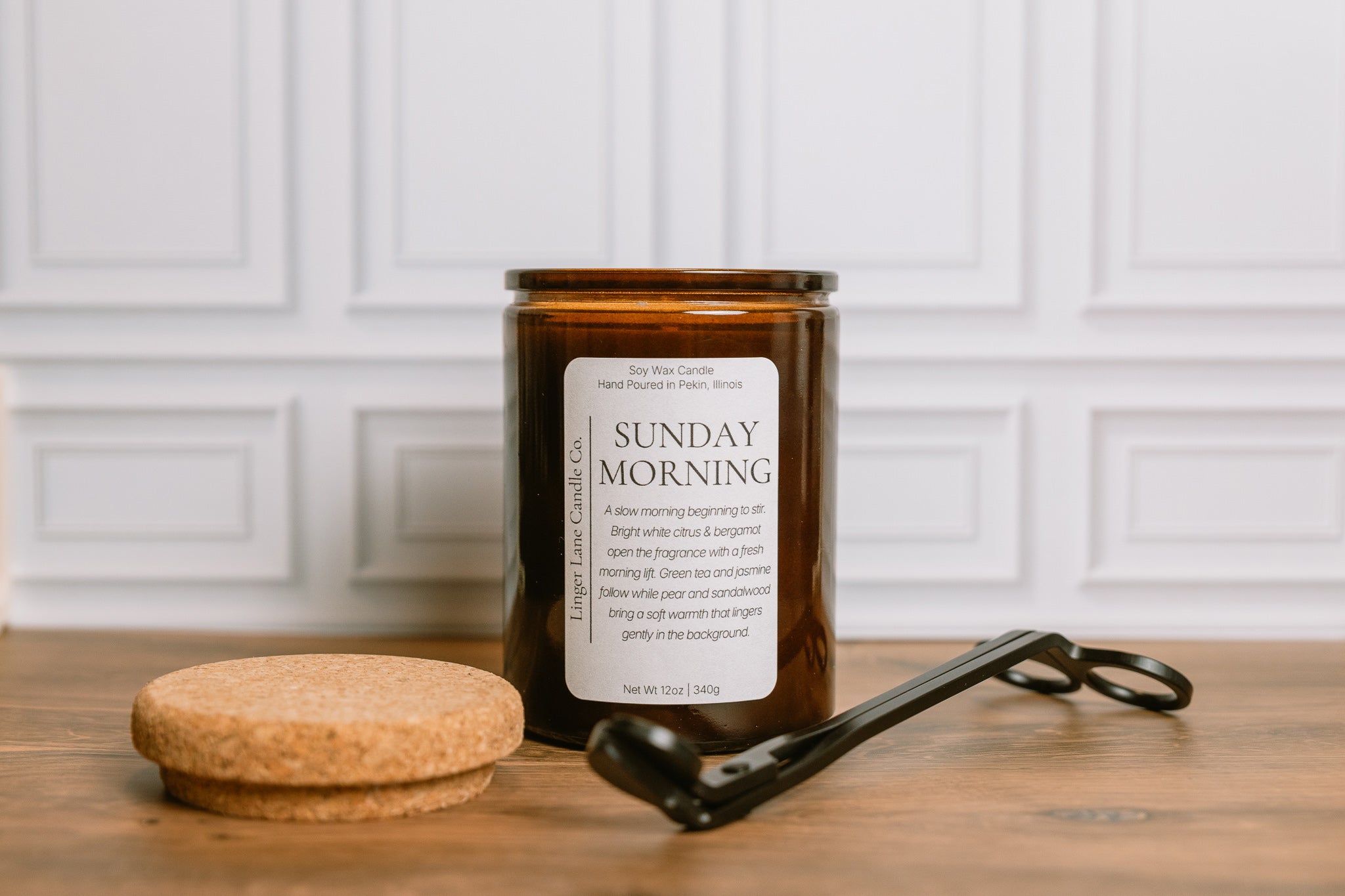 Home fragrance Candle labeled 'Sunday Morning' with a cork lid and wick trimmer on a wooden surface.