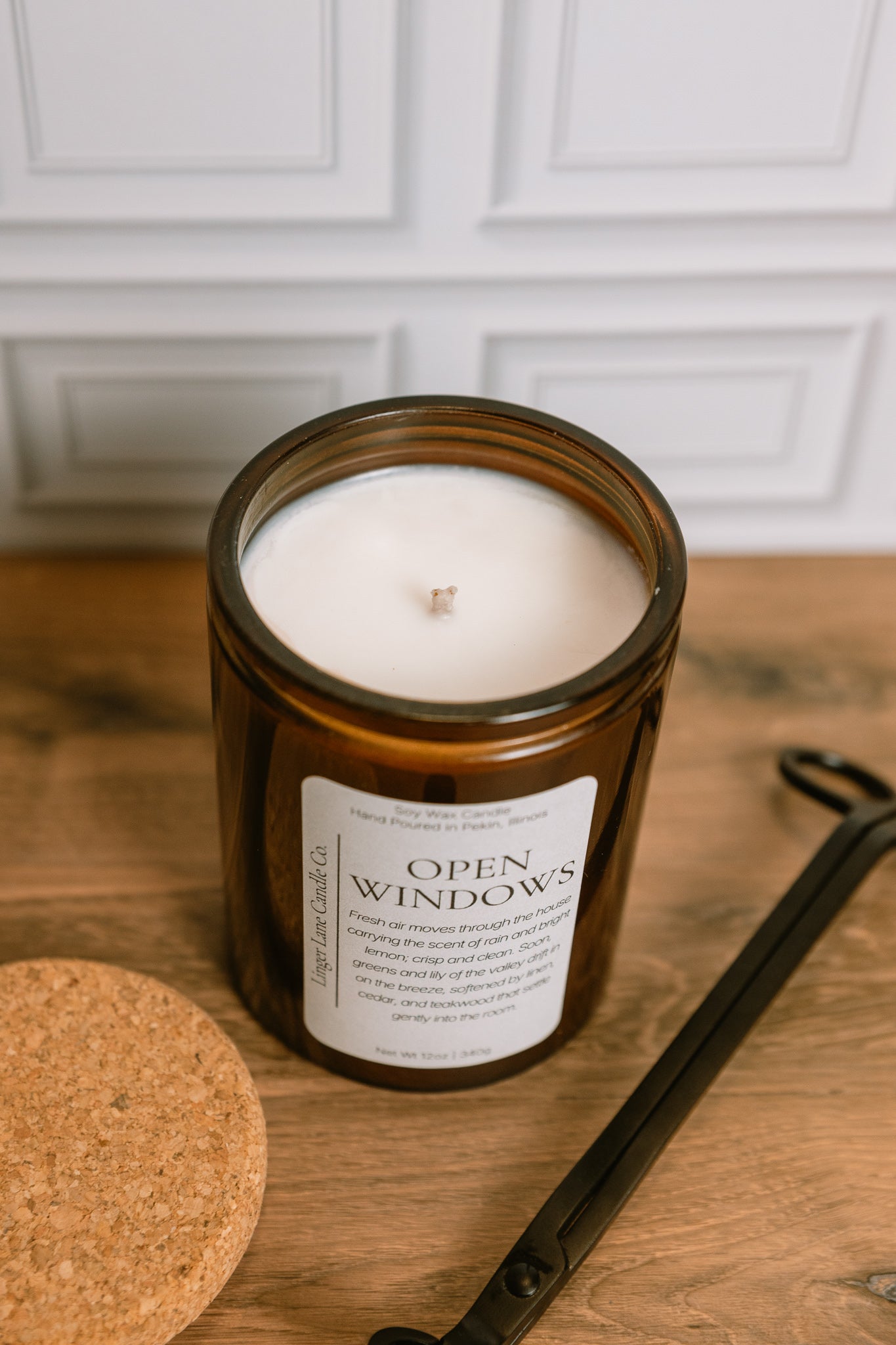 home fragrance white Candle in a dark amber colored glass jar labeled 'Open Windows' on a wooden surface with a white cabinet background.