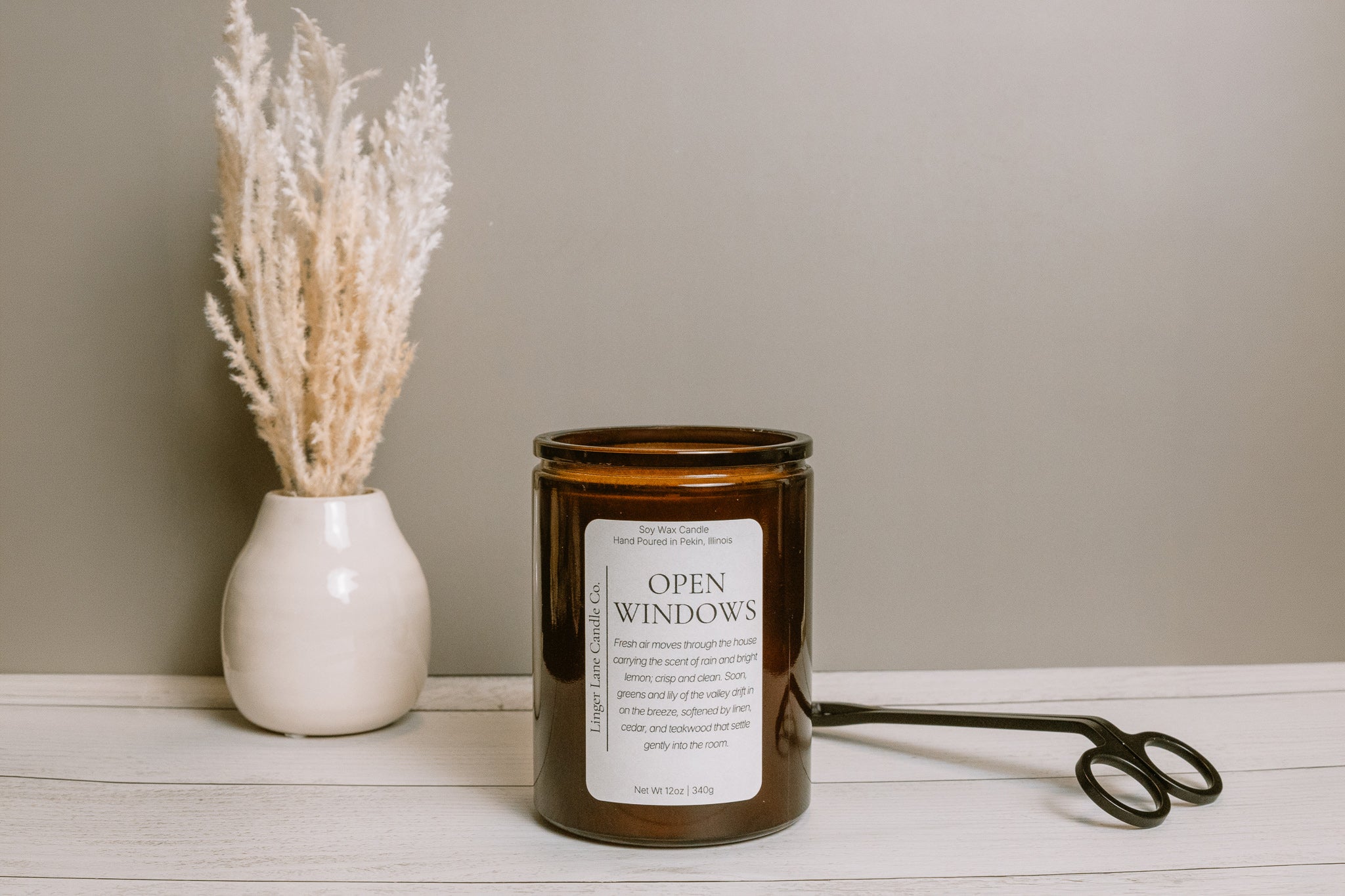 Soy wax candle in a dark amber colored candle jar labeled 'Open Windows' on a wooden surface with a vase of dried plants and scissors in the background.