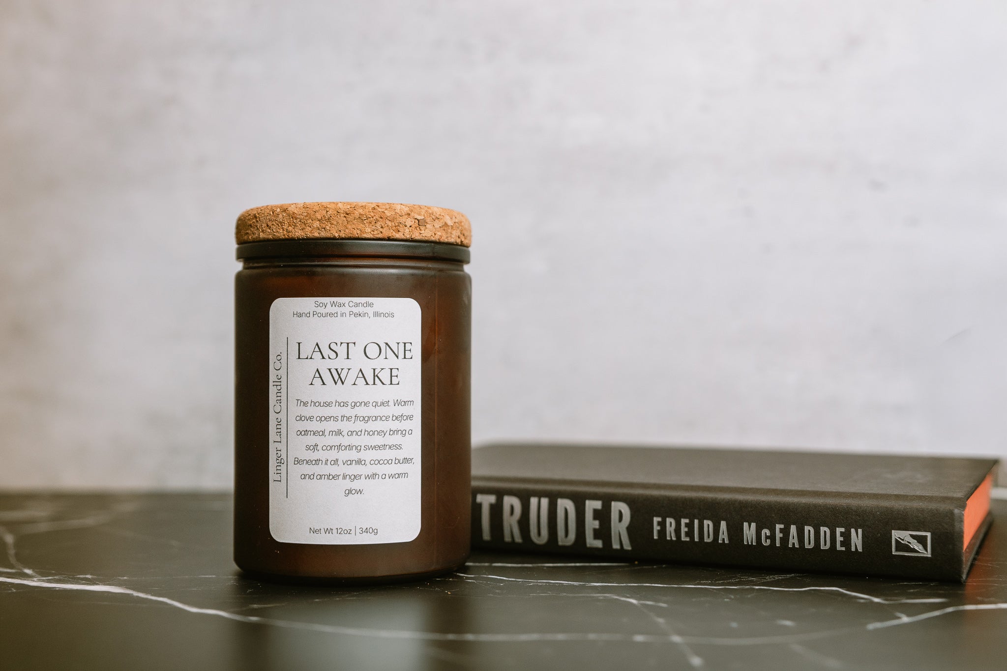 Frosted amber glass Candle jar with a soy wax candle labeled 'Last One Awake' on a surface with a book in the background