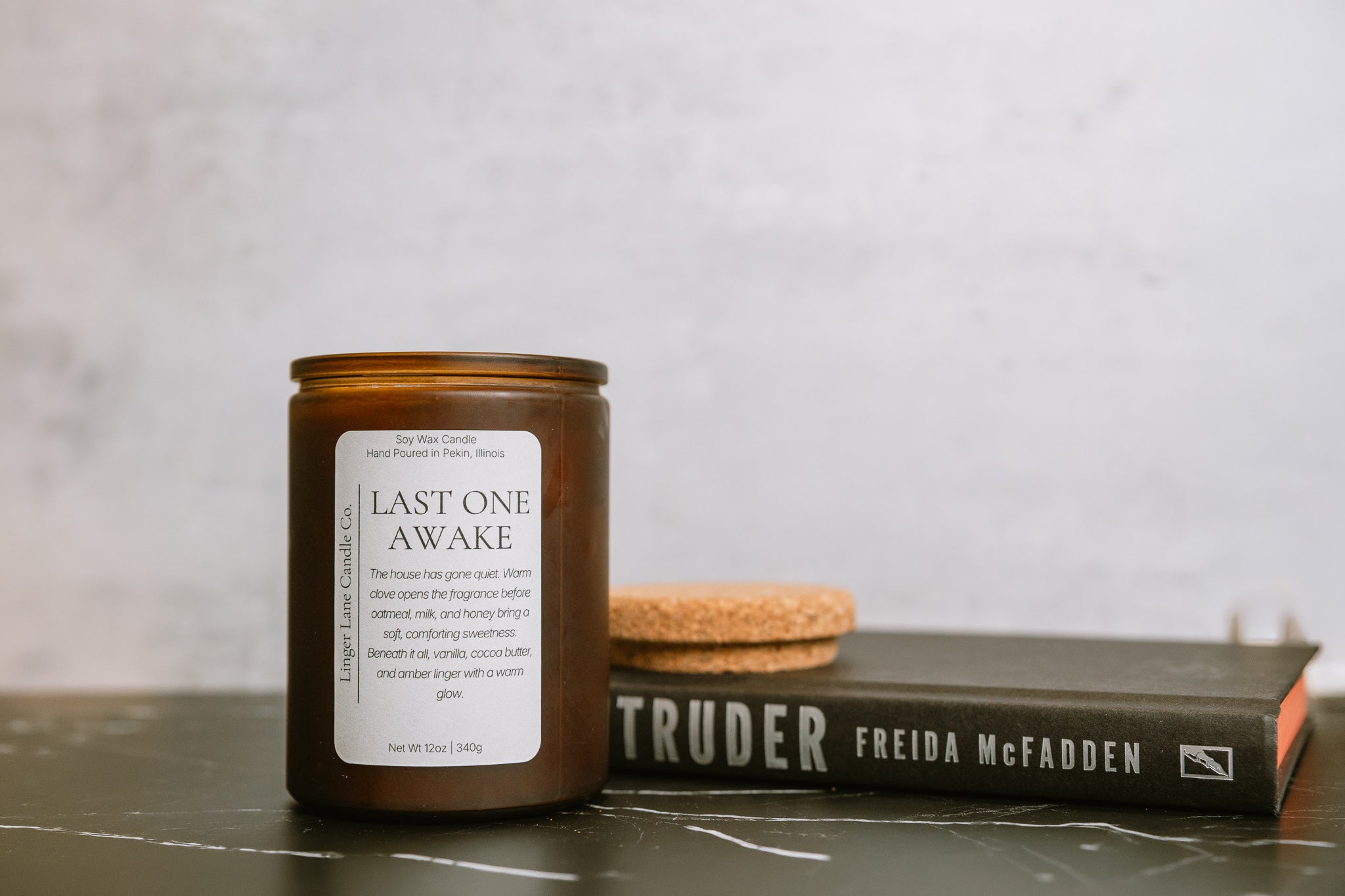 Soy wax Candle labeled 'Last One Awake' next to a book titled 'The Intruder' by Freida McFadden on a gray surface.