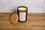 Scented soy wax Candle in a frosted amber glass jar with a cork lid on a wooden surface, featuring a label that reads 'Stay Awhile'.