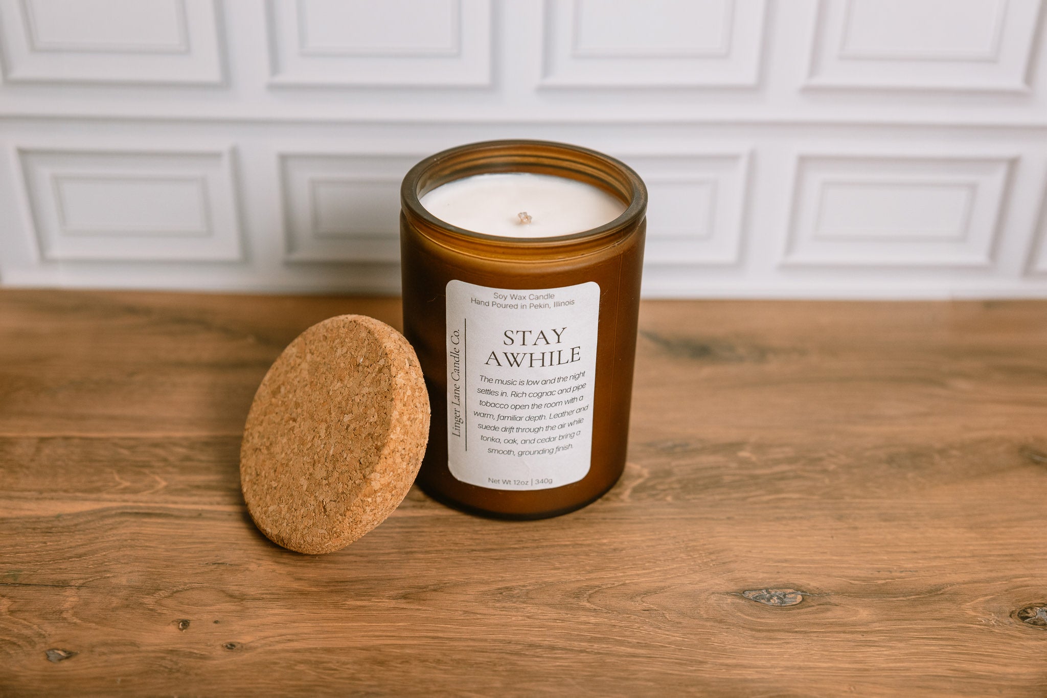 Scented soy wax Candle in a frosted amber glass jar with a cork lid on a wooden surface, featuring a label that reads 'Stay Awhile'.