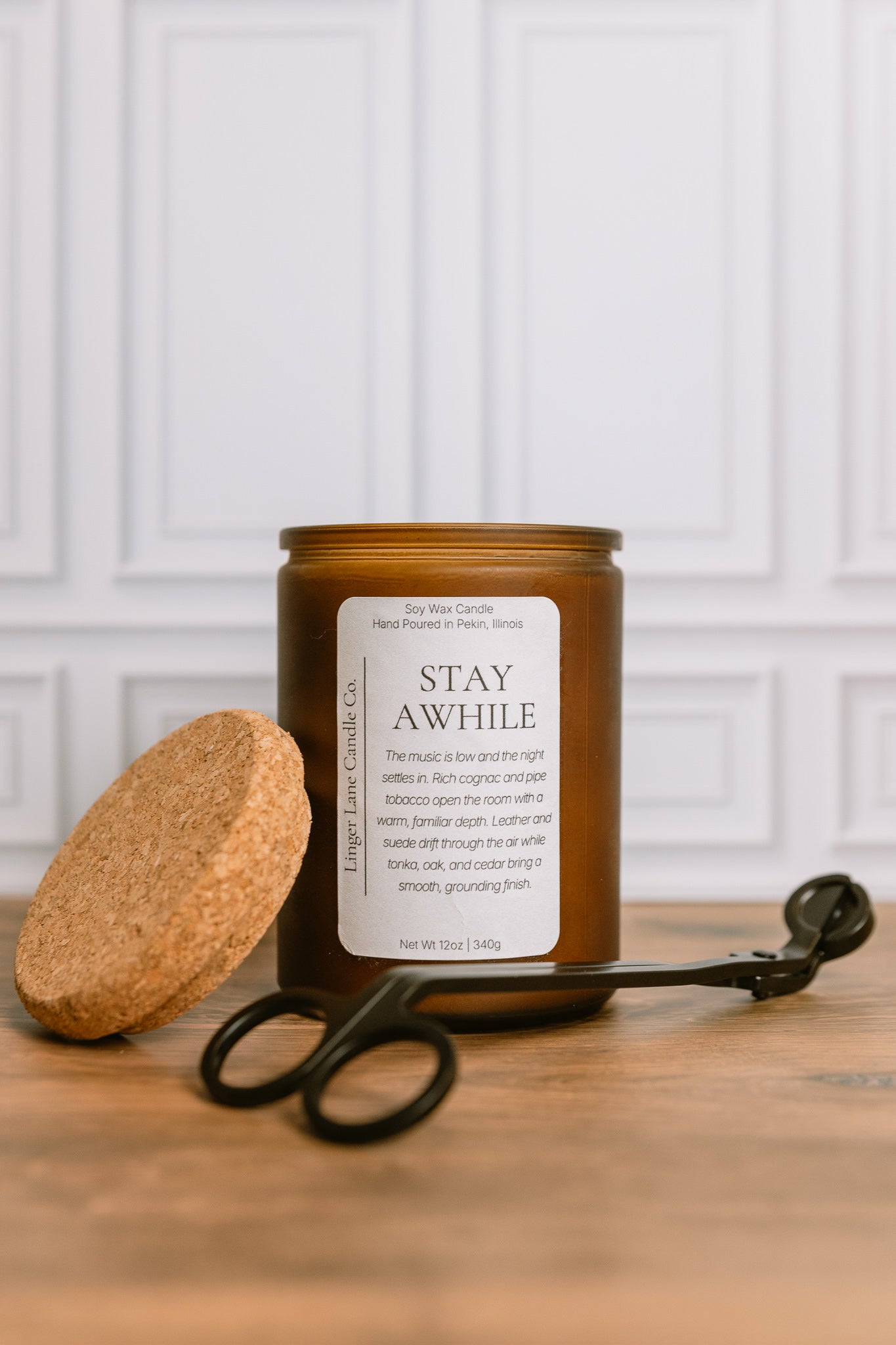 Frosted amber recycled glass Candle jar  containing a scented soy wax candle labeled 'Stay awhile' with a cork lid and black wick trimmer on a wooden surface.