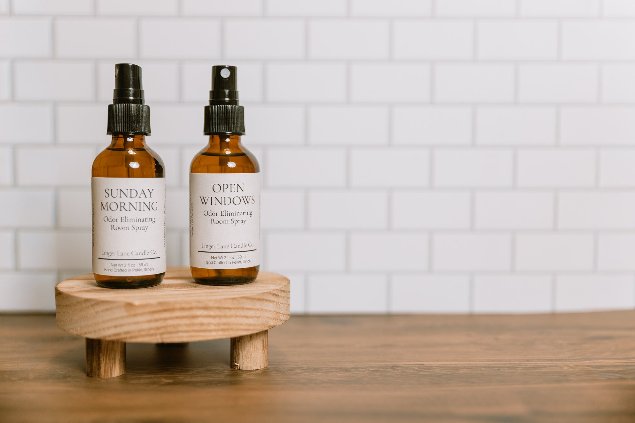 Two bottles of 'Sunday Morning' and 'Open Windows' odor eliminating room spray on a wooden stand with a tiled wall background.