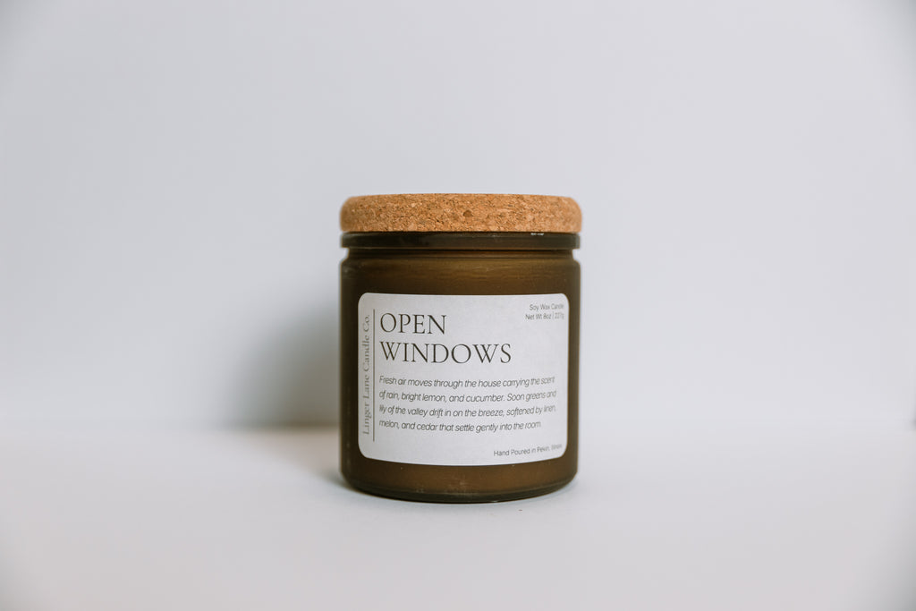 Scented soy Candle in a frosted amber jar labeled 'Open Windows' with a cork lid on a light gray background