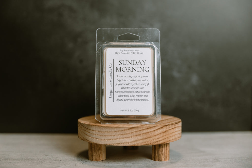 Packaged wax melt labeled 'Sunday Morning' on a wooden stand with a dark background